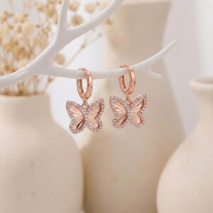 May include: Rose gold butterfly hoop earrings with clear crystal accents. The butterfly pendants are detailed with intricate wing designs and are suspended from small, round hoops. The earrings are displayed against a soft, neutral background.