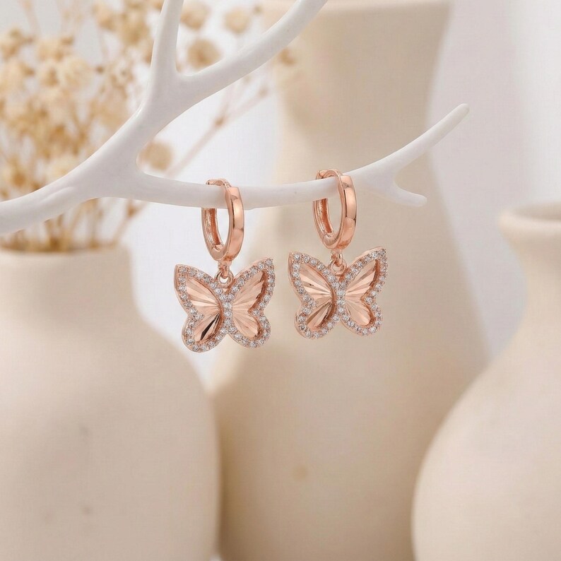May include: Rose gold butterfly hoop earrings with clear crystal accents. The butterfly pendants are detailed with intricate wing designs and are suspended from small, round hoops. The earrings are displayed against a soft, neutral background.