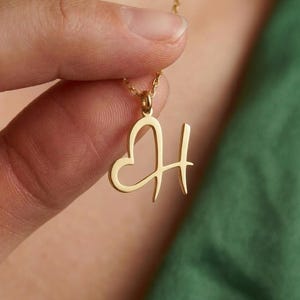 May include: A gold-colored pendant necklace featuring the letter "H" with a heart-shaped design. The pendant hangs from a delicate gold chain. The necklace is held against a green background.