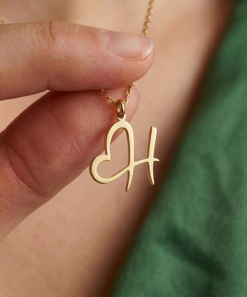 May include: A gold-colored pendant necklace featuring the letter "H" with a heart-shaped design. The pendant hangs from a delicate gold chain. The necklace is held against a green background.