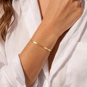 May include: A thin, polished gold-tone metal cuff bracelet with a smooth, flat surface and a minimalist, open-ended design. The bracelet features a simple, uniform band that encircles the wrist, showcasing a bright, reflective metallic finish.