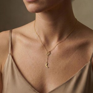 May include: A gold-colored necklace featuring a sun and crescent moon pendant. The sun pendant is at the top, with the moon pendant suspended below. The necklace is worn on a person's chest, with a neutral background.