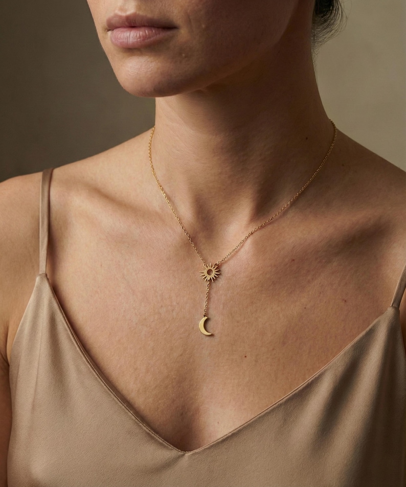 May include: A gold-colored necklace featuring a sun and crescent moon pendant. The sun pendant is at the top, with the moon pendant suspended below. The necklace is worn on a person's chest, with a neutral background.