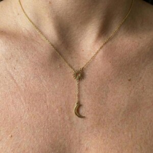 May include: A gold-colored necklace featuring a sun pendant connected to a chain that leads to a crescent moon charm. The necklace is worn around the neck, with the sun and moon pendants resting on the chest. The chain is delicate and the design is simple.