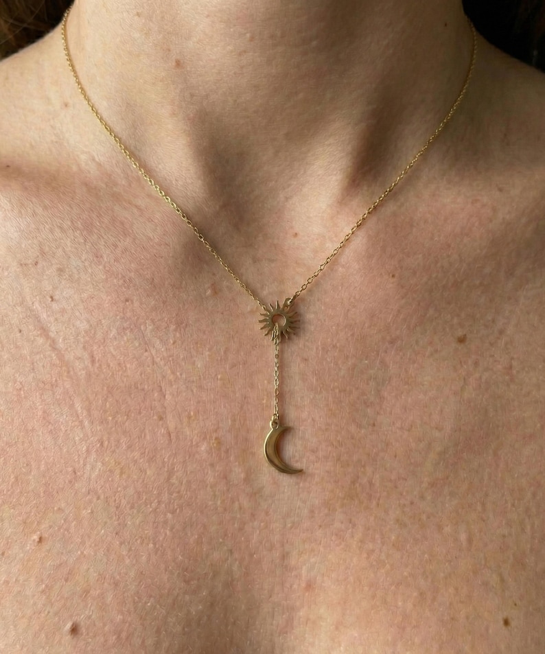 May include: A gold-colored necklace featuring a sun pendant connected to a chain that leads to a crescent moon charm. The necklace is worn around the neck, with the sun and moon pendants resting on the chest. The chain is delicate and the design is simple.