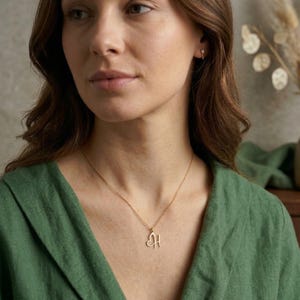 May include: A gold-colored necklace with a heart-shaped pendant featuring the letter "H". The necklace is worn against a green top. The woman in the image is wearing a small earring.