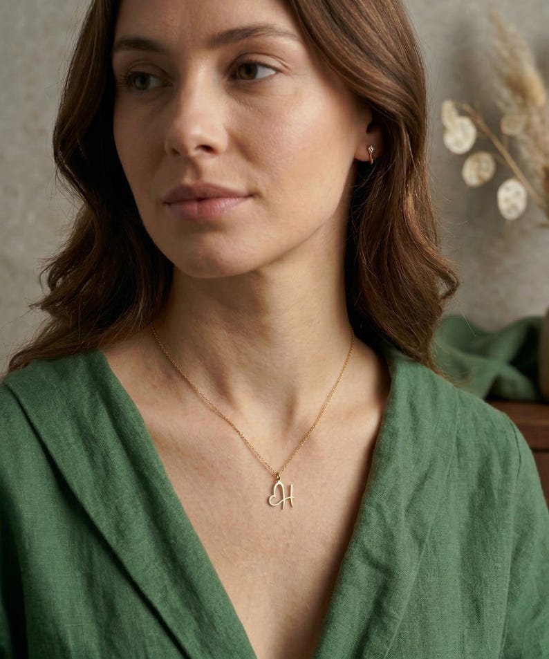 May include: A gold-colored necklace with a heart-shaped pendant featuring the letter "H". The necklace is worn against a green top. The woman in the image is wearing a small earring.
