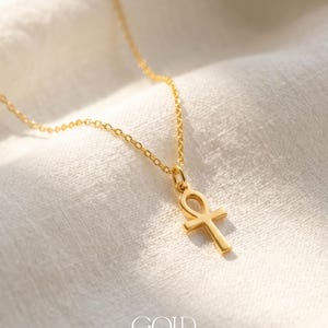 May include: A gold-colored necklace with an Ankh pendant, displayed on a white fabric background. The pendant is a cross-like shape with a loop at the top. The word "GOLD" is printed at the bottom of the image.