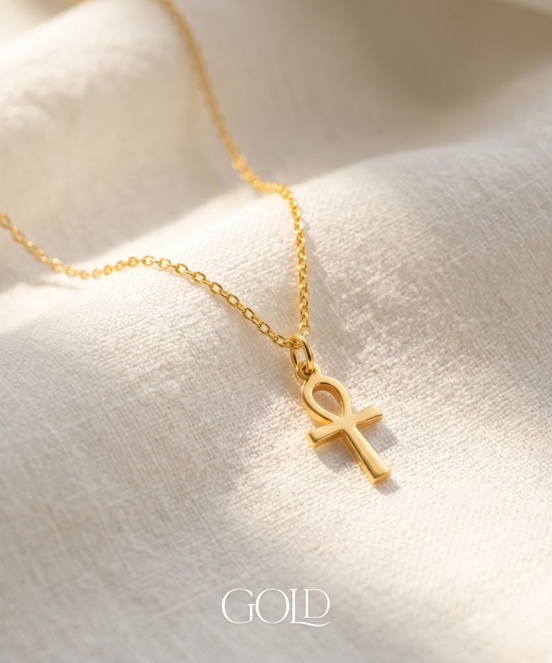 May include: A gold-colored necklace with an Ankh pendant, displayed on a white fabric background. The pendant is a cross-like shape with a loop at the top. The word "GOLD" is printed at the bottom of the image.