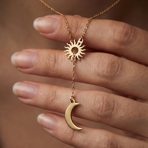 May include: A gold-colored necklace featuring a sun pendant at the top and a crescent moon pendant hanging below. The sun pendant has a detailed, radiant design, while the moon is a smooth, curved shape. The chain is delicate and links the two celestial charms.