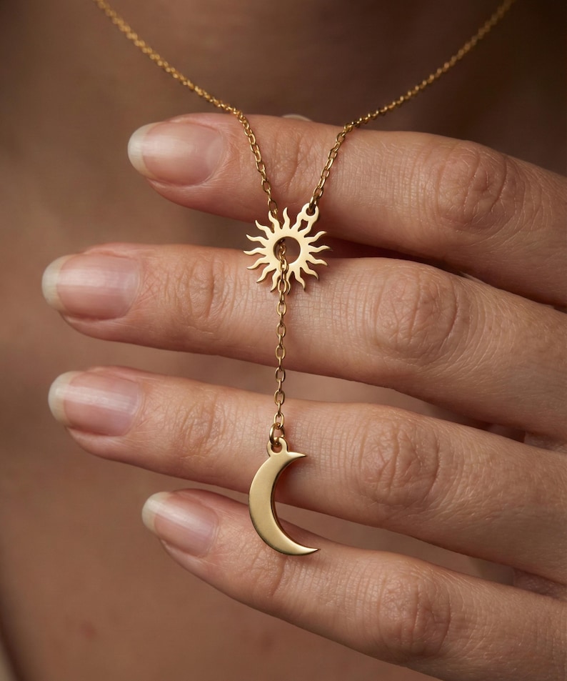 May include: A gold-colored necklace featuring a sun pendant at the top and a crescent moon pendant hanging below. The sun pendant has a detailed, radiant design, while the moon is a smooth, curved shape. The chain is delicate and links the two celestial charms.