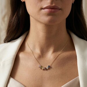 May include: A gold necklace with a delicate chain and a series of colorful gemstone pendants. The gemstones include pink, yellow, green, red, and blue, each with a unique shape and sparkle. The necklace is worn against a white blazer.