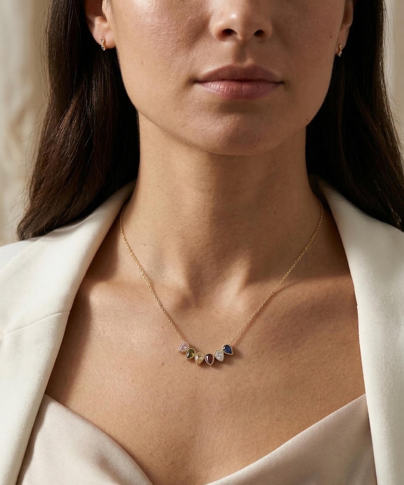 May include: A gold necklace with a delicate chain and a series of colorful gemstone pendants. The gemstones include pink, yellow, green, red, and blue, each with a unique shape and sparkle. The necklace is worn against a white blazer.