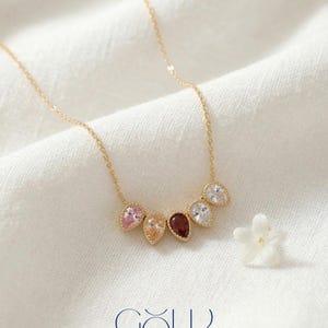 May include: A gold necklace with a delicate chain features six teardrop-shaped gemstones in various colors: pink, peach, dark red, and clear. The necklace is displayed on a white fabric with a small white flower and the word "GOLD" in blue.