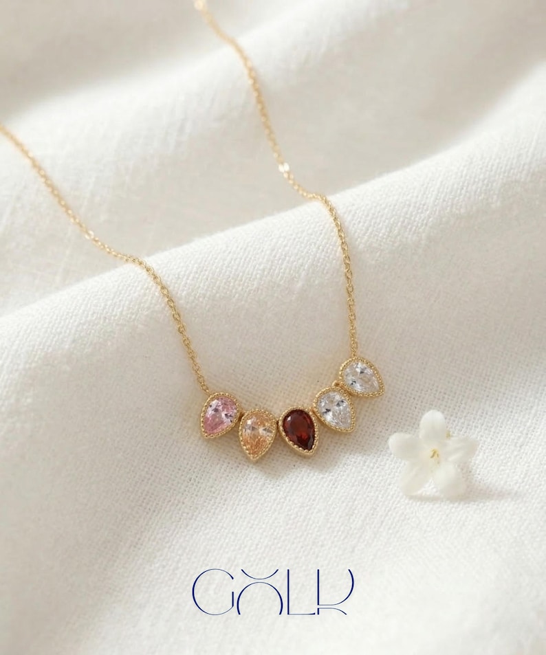 May include: A gold necklace with a delicate chain features six teardrop-shaped gemstones in various colors: pink, peach, dark red, and clear. The necklace is displayed on a white fabric with a small white flower and the word "GOLD" in blue.