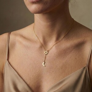 May include: A gold-colored necklace featuring a sun and crescent moon pendant. The sun pendant is at the top, with the moon pendant hanging below. The necklace is worn on a person wearing a gold-colored top.
