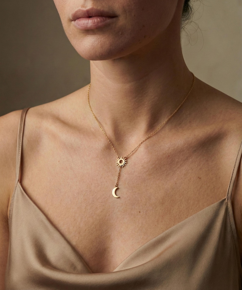 May include: A gold-colored necklace featuring a sun and crescent moon pendant. The sun pendant is at the top, with the moon pendant hanging below. The necklace is worn on a person wearing a gold-colored top.