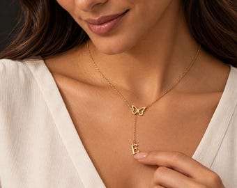 Pode incluir: Um colar lariat de tom dourado com uma corrente de elos finos e polidos, apresentando um pingente de borboleta delicado no centro e uma corrente que se estende para baixo até um pingente com a letra maiúscula E.