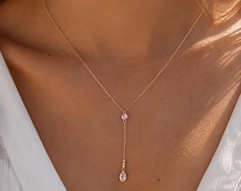 Pode incluir: Um colar tipo lariat em tom dourado com uma corrente fina, um pequeno detalhe de pedra preciosa rosa redonda e um pingente em forma de gota rosa pendurado na base, repousando sobre a clavícula.