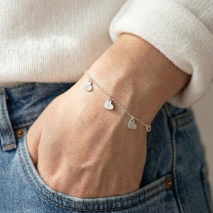 May include: A silver bracelet with three heart-shaped charms, each bearing a single initial. The bracelet is worn on a wrist, with the hand resting in a blue jean pocket. The person is wearing a white sweater.