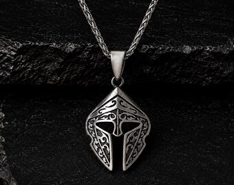 Pode incluir: Colar com pingente prateado com um design detalhado de capacete espartano. O pingente é intrincadamente esculpido com padrões ornamentais e pende de uma corrente a condizer. O colar é exibido contra um fundo escuro e texturizado.