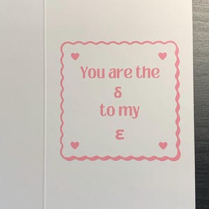 Może przedstawiać: Biała kartka z życzeniami z różową, falistą obwódką i różowym napisem "You are the δ to my ε". Małe różowe serca znajdują się w każdym rogu obwódki. Karta jest otwarta, ukazując wnętrze.