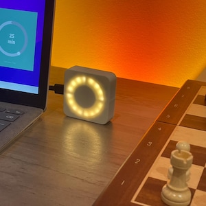 Könnte beinhalten: Ein weißer, quadratischer Timer mit kreisförmiger Lichtanzeige ist an einen Laptop angeschlossen. Der Laptop-Bildschirm zeigt einen Timer, der auf 25 Minuten eingestellt ist. Ein Schachbrett mit schwarzen und weißen Figuren befindet sich im Hintergrund.