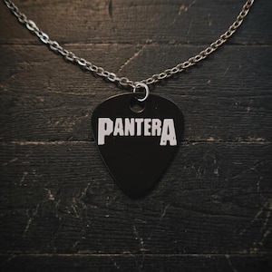 May include: A black guitar pick pendant with the word "PANTERA" in white block letters. The pendant hangs from a silver chain. The background is a dark, textured wooden surface.