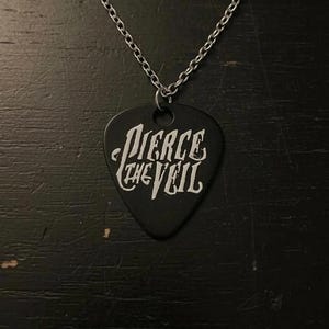 Puede incluir: Un collar con colgante de púa de guitarra negra con la inscripción blanca "Pierce the Veil". El colgante tiene forma de púa de guitarra y cuelga de una cadena plateada. El fondo es una superficie oscura y texturizada.