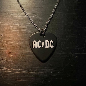 May include: Black guitar pick pendant necklace with the AC/DC logo in white. The pendant hangs from a silver-colored chain. The necklace is displayed on a dark, textured surface.
