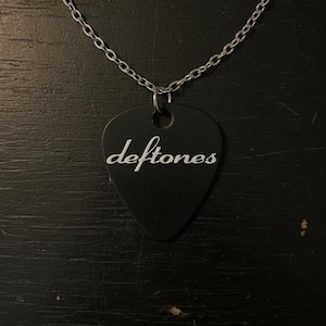 Peut inclure: Un collier avec un pendentif en forme de médiator noir portant l'inscription "deftones" en blanc. Le pendentif est suspendu à une chaîne argentée. Le collier est présenté sur un fond sombre.