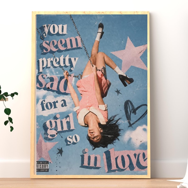 May include: A framed wall art print featuring a person hanging upside down from a swing against a blue sky background. The text reads, "you seem pretty sad for a girl so in love." The design includes pink stars, a hand-drawn heart, and a black label.