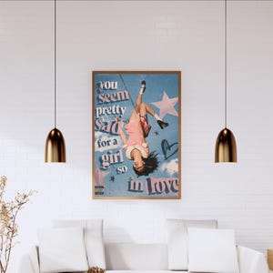 May include: A rectangular wall poster featuring a person in a pink dress hanging upside down from a chain against a blue sky with clouds and stars. The text reads, "you seem pretty sad for a girl so in love," with a small parental advisory label.