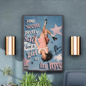 May include: A rectangular wall art print featuring a person hanging upside down from a swing against a blue sky background with pink stars and a heart. The text reads: you seem pretty sad for a girl so in love. A small parental advisory label is visible.