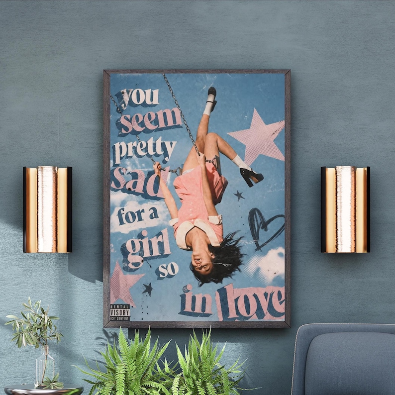 May include: A rectangular wall art print featuring a person hanging upside down from a swing against a blue sky background with pink stars and a heart. The text reads: you seem pretty sad for a girl so in love. A small parental advisory label is visible.