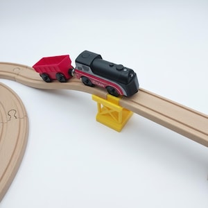 Könnte beinhalten: Ein hölzernes Spielzeug-Zugset mit einer schwarz-roten Lokomotive und einem roten Güterwagen. Der Zug fährt auf einer Holzstrecke über eine gelbe Brücke. Das Set ist für Kinderspiele konzipiert.