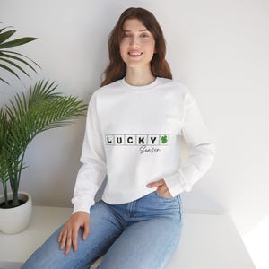 Puede incluir: Sudadera blanca con el texto "LUCKY Season" en negro y verde. La palabra "LUCKY" está escrita con fichas tipo Scrabble. Un trébol verde está al lado de la palabra "LUCKY". La sudadera es de manga larga.