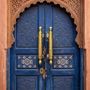 May include: A detailed shot of a blue wooden door with intricate geometric carvings and gold-colored handles. The door features a mosaic tile design and is framed by a carved wooden archway, set against a reddish-brown wall.