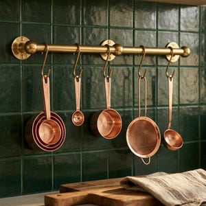 May include: A horizontal brass kitchen rail mounted on a wall, featuring five S-shaped hooks. Hanging from these hooks are a set of nested copper measuring cups, a small copper measuring spoon, a copper measuring cup, a copper sieve, and a ladle.