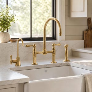 May include: A polished brass kitchen tap set featuring a high-arc bridge tap with two lever handles, a separate side spray, and a smaller filtered water dispenser, all mounted on a white marble worktop above a white farmhouse sink.