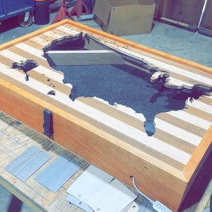Puede incluir: Una caja rectangular de madera con una superficie superior de franjas de madera claras y oscuras alternadas. Un recorte con la forma de los Estados Unidos está relleno de resina epoxi negra. Un pestillo negro está en el lateral y un cable blanco.
