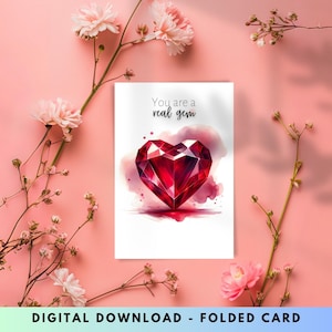 Peut inclure: Une carte pliée avec une illustration à l'aquarelle d'un joyau rouge en forme de cœur. Le texte "You are a real gem" est au-dessus du joyau. La carte est entourée de fleurs et de branches roses sur un fond rose.