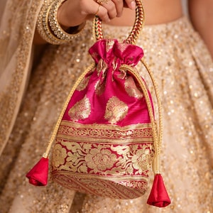 Może przedstawiać: Żywa różowa torba ze sznurkiem ze złotymi kwiatowymi wzorami i wykończeniami. Torba ma pleciony uchwyt i pasujące frędzle. Tło przedstawia złotą, zdobioną tkaninę, sugerującą uroczystą lub formalną okazję.