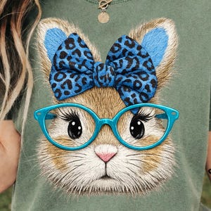 May include: Olive green t-shirt featuring a whimsical illustration of a cat's face. The cat wears blue glasses and a blue bow with a leopard print. The cat's ears are also blue. The design is centered on the shirt.