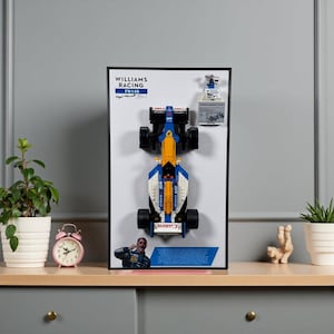 May include: Framed Williams Racing FW14B model car. The model is primarily blue, yellow, and white, with black wheels. The frame includes the Williams Racing logo, the FW14B model name, and a photo of Nigel Mansell. A small trophy and a diagram are also included.