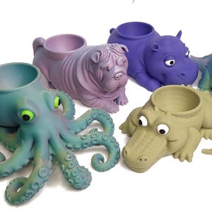 May include: A selection of animal-shaped plant pots. The pots feature an octopus, a Shar Pei dog, a hippo, a triceratops, and an alligator. Each pot has a circular opening for plants and is painted in different colours.
