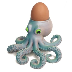 May include: A novelty egg cup in the shape of an octopus, in blue and purple hues, with vivid green eyes. It holds a brown egg. The tentacles feature suction cup detailing. A quirky kitchen accessory.