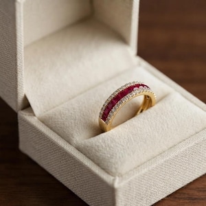 Op de afbeelding: Een gouden ring met een centrale rij vierkant geslepen rode robijnen, geflankeerd door twee rijen kleine ronde witte diamanten, gepresenteerd in een crèmekleurig met stof bekleed juwelendoosje met een getextureerde buitenkant.