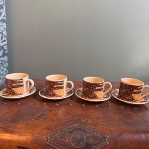 Può includere: Un set di quattro tazzine da caffè in ceramica color crema con piattini abbinati. Ogni tazzina presenta una stampa grafica marrone di chicchi di caffè e la parola COFFEE in carattere scuro. I piattini hanno un motivo circolare scuro.
