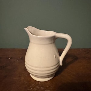 May include: A small, off-white ceramic pitcher with a curved handle and a spout. The pitcher has a rounded body with horizontal ridges near the base. The background is a solid teal color, and the pitcher is sitting on a wooden surface.
